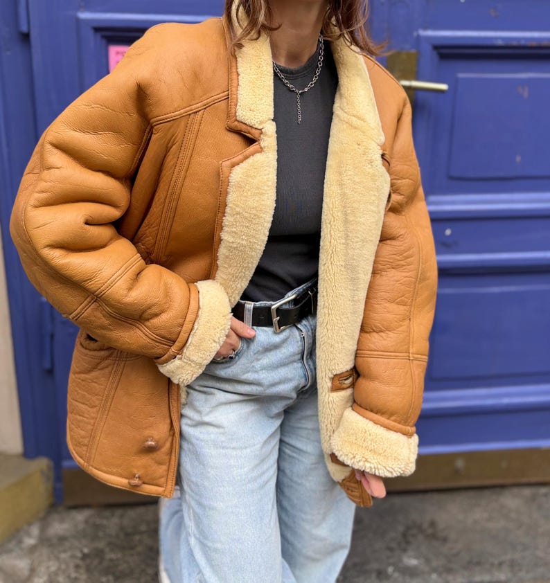 May include: A brown shearling jacket with a cream-colored shearling lining and trim. The jacket has a classic lapel collar, button closures, and a belted waist. The jacket is worn with a black belt and light blue jeans.
