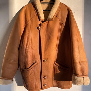 May include: A brown leather coat with a beige shearling collar and cuffs. The coat has a button-up front, two front pockets, and decorative stitching. The coat is hanging on a black hanger.