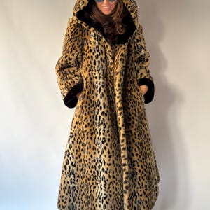 May include: A long, hooded faux fur coat featuring a tan and black leopard print pattern. The coat includes a deep hood, long sleeves with black cuffs, and a black interior lining that is visible along the front opening and the hood edge.
