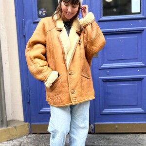 May include: A tan shearling coat with a cream-colored interior and a button-up front. The coat has a classic design with a notched collar and pockets. The person is wearing a gray beanie, light blue jeans, and white sneakers.
