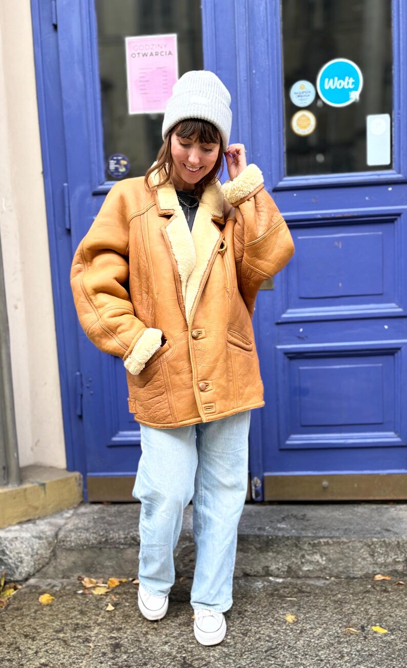 May include: A tan shearling coat with a cream-colored interior and a button-up front. The coat has a classic design with a notched collar and pockets. The person is wearing a gray beanie, light blue jeans, and white sneakers.