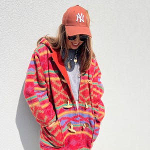 May include: A person wearing a rust-colored baseball cap with a white "NY" logo, sunglasses, and a red, orange, and blue patterned hooded coat with wooden toggles. The coat is zipped up and has pockets.