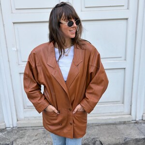 May include: A cognac-coloured leather blazer featuring a double-breasted front with two visible buttons, wide lapels, and long sleeves. The jacket includes two front pockets and is worn over a plain white top and light blue denim jeans.