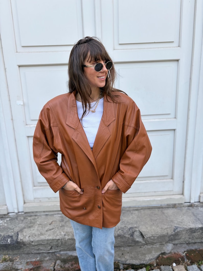 May include: A cognac-coloured leather blazer featuring a double-breasted front with two visible buttons, wide lapels, and long sleeves. The jacket includes two front pockets and is worn over a plain white top and light blue denim jeans.