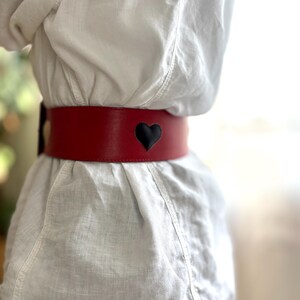 May include: A wide, red leather belt with a black heart-shaped cutout. The belt is worn around the waist and is the focal point of the image. The belt is approximately 3 inches wide.