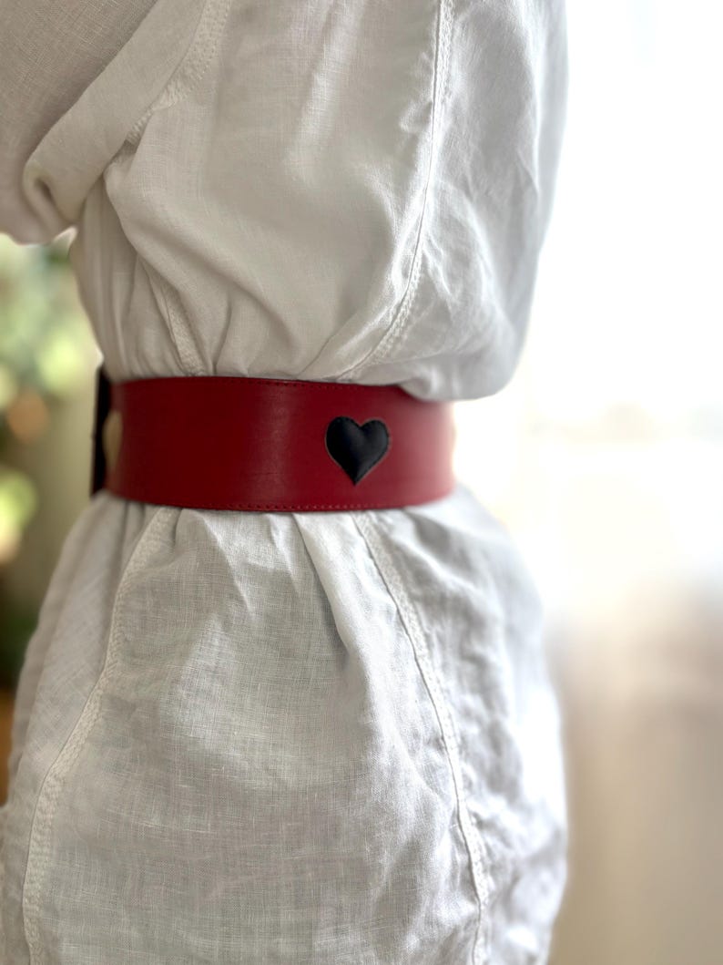 May include: A wide, red leather belt with a black heart-shaped cutout. The belt is worn around the waist and is the focal point of the image. The belt is approximately 3 inches wide.