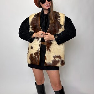 May include: A vintage-inspired outfit featuring a brown and white cowhide vest over a black top and skirt. The ensemble includes a brown beret, large sunglasses, and black knee-high boots. The vest is the focal point.