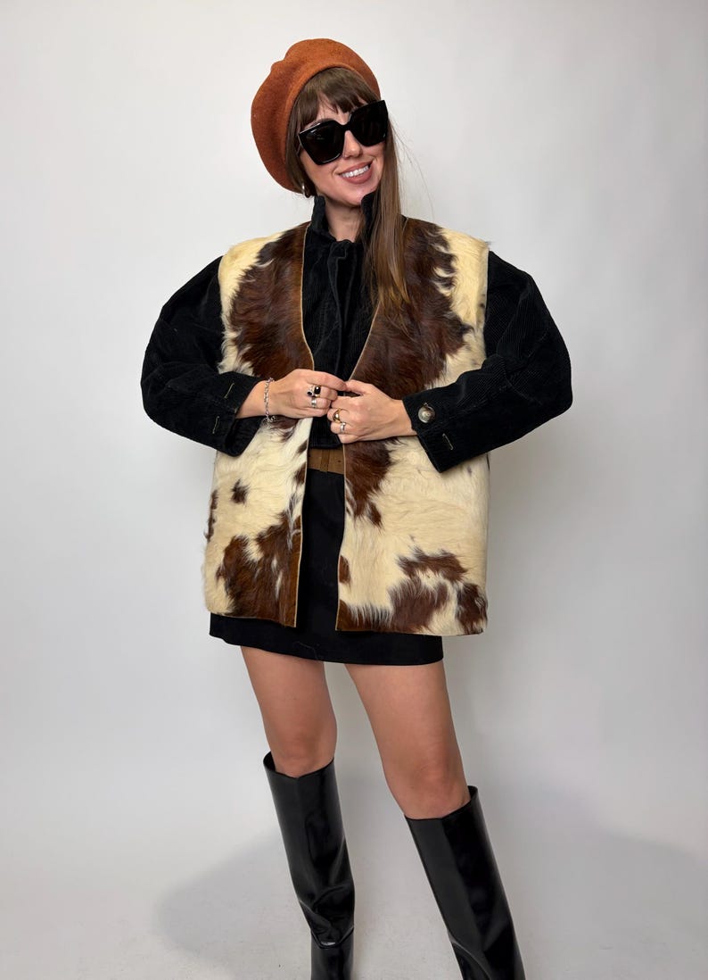 May include: A vintage-inspired outfit featuring a brown and white cowhide vest over a black top and skirt. The ensemble includes a brown beret, large sunglasses, and black knee-high boots. The vest is the focal point.