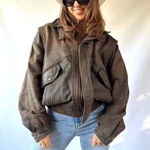Pode incluir: Uma jaqueta bomber de couro marrom escuro com gola, mangas compridas e dois bolsos frontais com aba e fecho de pressão. A jaqueta é combinada com jeans azul claro, um chapéu de cowboy branco e óculos de sol pretos com lentes ovais.