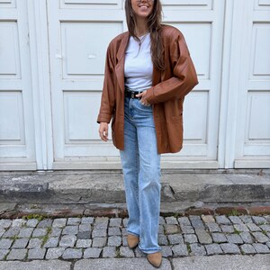 May include: A brown leather blazer worn over a white crew neck t-shirt and light blue denim jeans. The outfit includes a black belt with a silver buckle, round sunglasses, and tan pointed-toe shoes. The blazer features long sleeves and pockets.