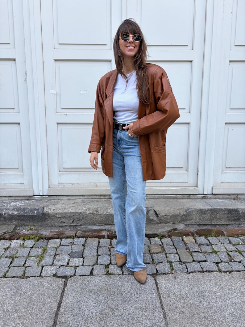 May include: A brown leather blazer worn over a white crew neck t-shirt and light blue denim jeans. The outfit includes a black belt with a silver buckle, round sunglasses, and tan pointed-toe shoes. The blazer features long sleeves and pockets.