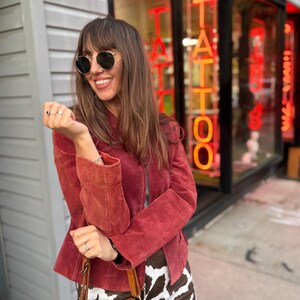May include: A woman wearing a red suede jacket, round sunglasses, and a cow print skirt. She is holding a brown leather bucket bag. The background features a tattoo parlor with neon signs.