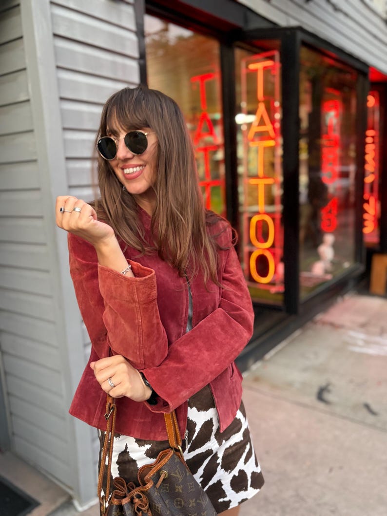 May include: A woman wearing a red suede jacket, round sunglasses, and a cow print skirt. She is holding a brown leather bucket bag. The background features a tattoo parlor with neon signs.