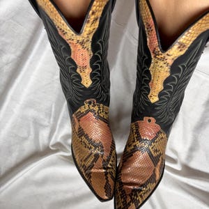 Puede incluir: Un par de botas de vaquero negras con un estampado de piel de serpiente en la puntera y la parte superior de la ca&ntilde;a. Las botas tienen una puntera puntiaguda y costuras decorativas. El estampado de piel de serpiente presenta tonos marrones, beiges y rosas.