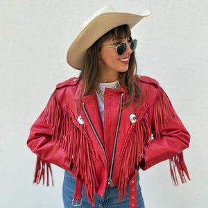 Op de afbeelding: Een rode leren jas met lange franjes en een zilveren ritssluiting. De jas heeft gevlochten accenten en een bijpassende rode riem. De persoon draagt een cr&egrave;mekleurige cowboyhoed en een zonnebril.