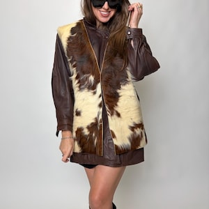 May include: A brown leather jacket with a cowhide vest featuring brown and cream patches. The outfit includes a brown beret and black sunglasses. The overall style is vintage and fashionable.