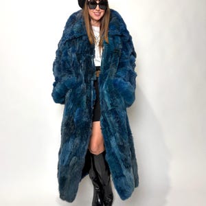 May include: A long, vibrant blue fur coat with a textured appearance. The coat has a wide collar and long sleeves. The person is wearing a black beret, sunglasses, and black boots. The coat is the focal point of the image.