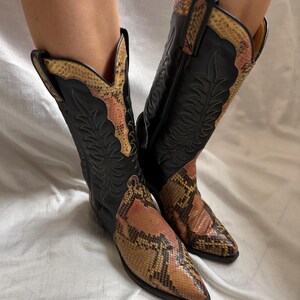 Puede incluir: Un par de botas de vaquero negras con un estampado de piel de serpiente en el pie y el borde superior. Las botas tienen una punta puntiaguda y costuras decorativas. El estampado de piel de serpiente presenta tonos marrones, beiges y rosas.