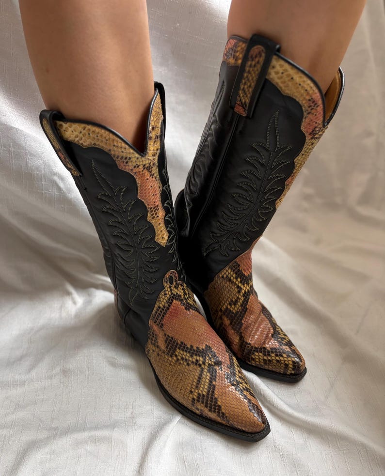 Puede incluir: Un par de botas de vaquero negras con un estampado de piel de serpiente en el pie y el borde superior. Las botas tienen una punta puntiaguda y costuras decorativas. El estampado de piel de serpiente presenta tonos marrones, beiges y rosas.