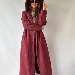 May include: A long, deep burgundy coat featuring a textured, fleece-like material, a large hood, and a front button closure. The coat extends to the lower calf and has long sleeves, providing a full-length silhouette with a consistent dark red colour.