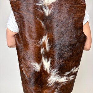 May include: A brown and white cowhide vest. The vest is sleeveless and has a natural, textured appearance. The color is a mix of dark brown and white patches, creating a unique pattern. The vest is shown from the back.