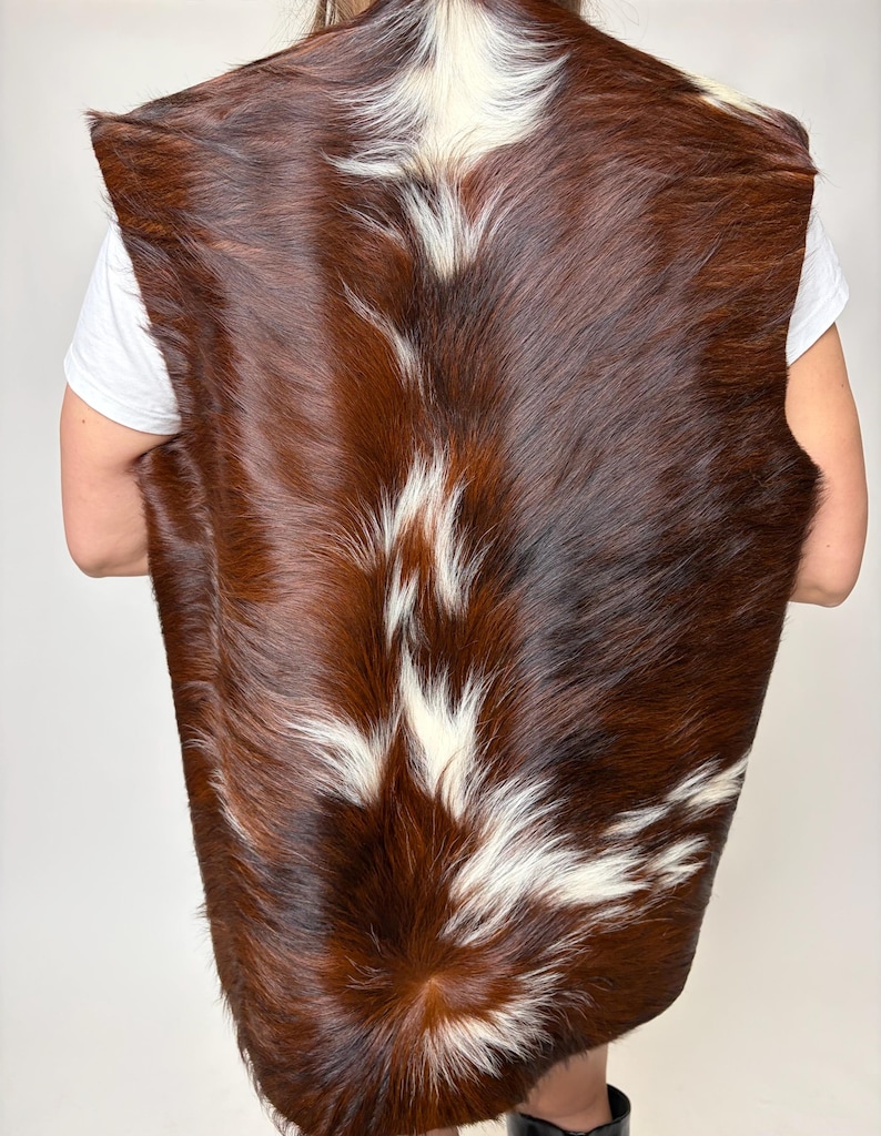 May include: A brown and white cowhide vest. The vest is sleeveless and has a natural, textured appearance. The color is a mix of dark brown and white patches, creating a unique pattern. The vest is shown from the back.