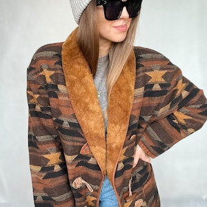 Peut inclure: Un manteau à motifs marrons et noirs avec un col en fausse fourrure marron et des fermetures à boutons. Le manteau est porté sur un pull gris et un jean bleu. La personne porte un bonnet gris et des lunettes de soleil noires.