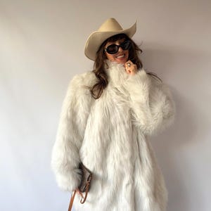 May include: A long-sleeved, off-white faux fur coat with a high collar, paired with a beige cowboy hat, dark oval sunglasses, and a small brown leather handbag. The coat features a thick, shaggy texture and reaches down to the hips.