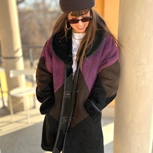 Può includere: Un cappotto a maniche lunghe caratterizzato da un design a blocchi di colore con pannelli viola, marroni e neri. Il cappotto presenta un ampio colletto e bordi in ecopelliccia nera, chiusura frontale con bottoni e una consistenza morbida.