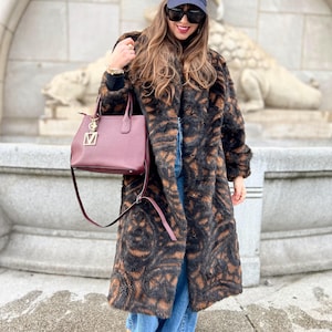 May include: A long, faux fur coat with a brown and black animal print. The coat is paired with blue jeans, a burgundy handbag, and a navy baseball cap with a white logo. The outfit is completed with sunglasses and tan shoes.