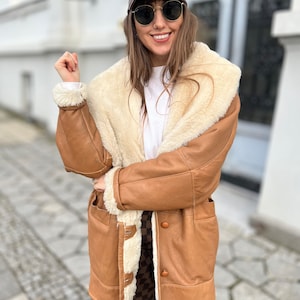 May include: A brown leather jacket with a white shearling lining. The jacket has a collar, a button closure, and two pockets. The person wearing the jacket is also wearing a brown baseball cap and sunglasses.
