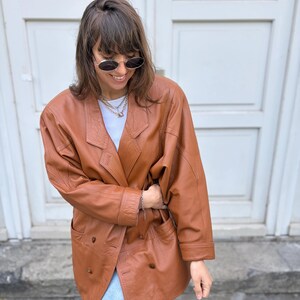May include: A double-breasted cognac brown leather blazer featuring wide lapels, long sleeves with cuffs, and two front pockets. The jacket is worn over a plain white t-shirt and paired with light blue denim jeans, accessorised with a layered chain necklace.