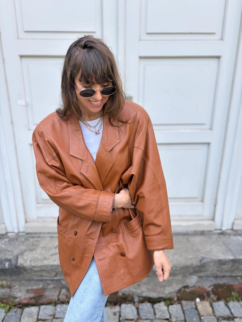 May include: A double-breasted cognac brown leather blazer featuring wide lapels, long sleeves with cuffs, and two front pockets. The jacket is worn over a plain white t-shirt and paired with light blue denim jeans, accessorised with a layered chain necklace.