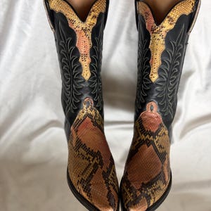 Puede incluir: Un par de botas de vaquero negras con un estampado de piel de serpiente en el pie y el cuello superior. Las botas tienen una punta puntiaguda y costuras decorativas. El estampado de piel de serpiente presenta tonos marrones, beige y rosa.