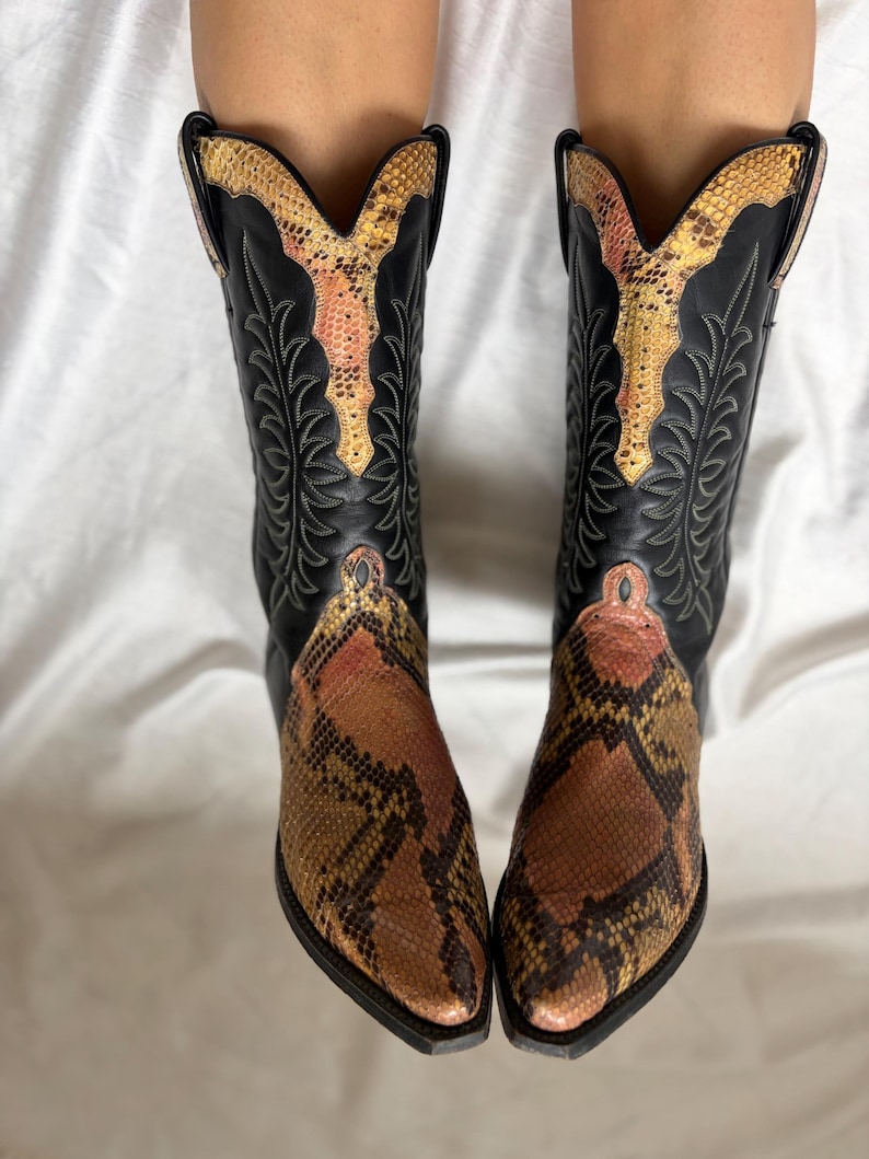 Puede incluir: Un par de botas de vaquero negras con un estampado de piel de serpiente en el pie y el cuello superior. Las botas tienen una punta puntiaguda y costuras decorativas. El estampado de piel de serpiente presenta tonos marrones, beige y rosa.