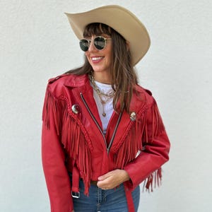 Op de afbeelding: Een rode leren jas met franjes, een zilveren rits en een riem. De jas wordt gecombineerd met een blauwe spijkerbroek en een beige cowboyhoed. De persoon draagt ook een zonnebril en een gouden ketting.