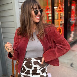 May include: A woman wearing a red suede jacket, gray top, and a cow print skirt. She is wearing sunglasses and smiling. A brown leather handbag is visible. The background includes a storefront with the word "TATTOO" in red neon.