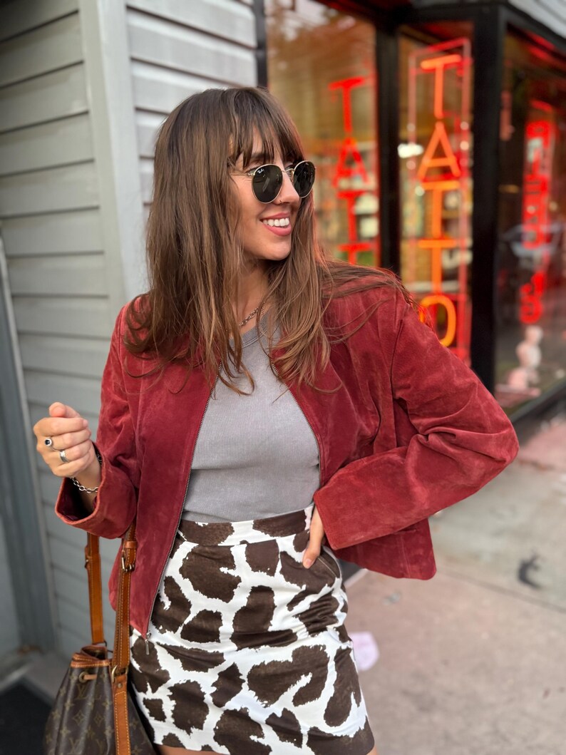 May include: A woman wearing a red suede jacket, gray top, and a cow print skirt. She is wearing sunglasses and smiling. A brown leather handbag is visible. The background includes a storefront with the word "TATTOO" in red neon.