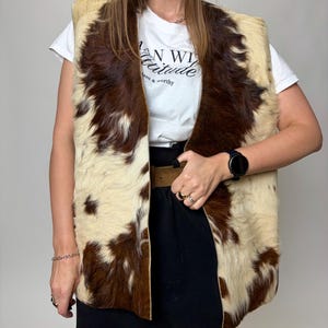 May include: A cowhide vest with a cream and brown pattern. The vest is open-fronted and sleeveless. The model is wearing a white t-shirt with the text "AN WILD attitude" and a black skirt.
