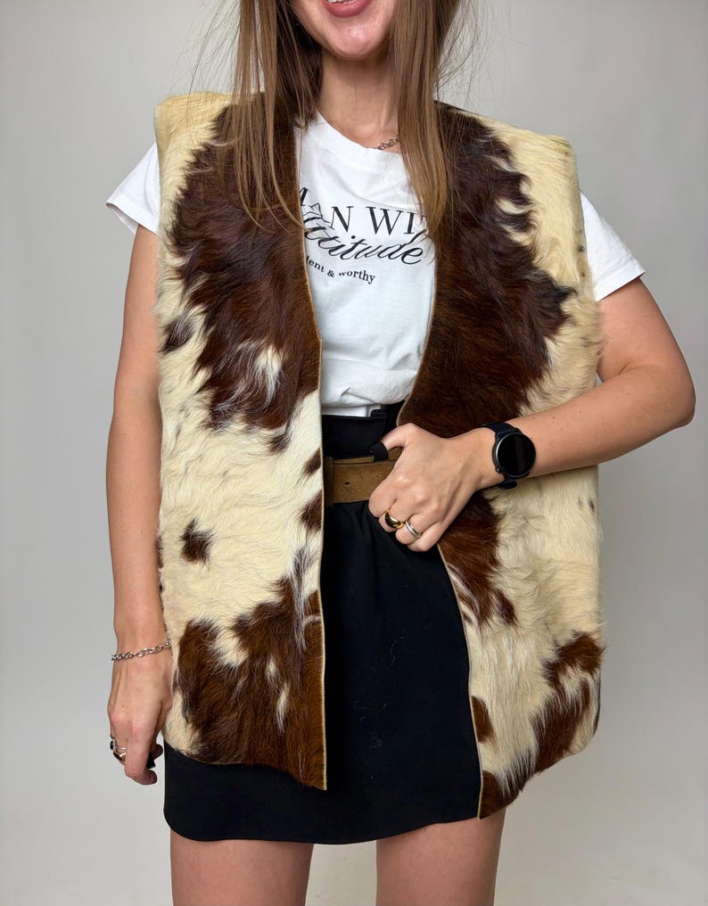 May include: A cowhide vest with a cream and brown pattern. The vest is open-fronted and sleeveless. The model is wearing a white t-shirt with the text "AN WILD attitude" and a black skirt.