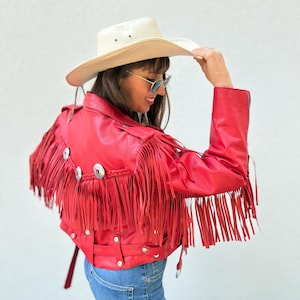 Op de afbeelding: Rode leren jas met lange franjes en zilverkleurige versieringen. De jas wordt gecombineerd met een blauwe spijkerbroek en een cr&egrave;mekleurige cowboyhoed. De franjes geven een Western-stijl. De jas heeft een riem.
