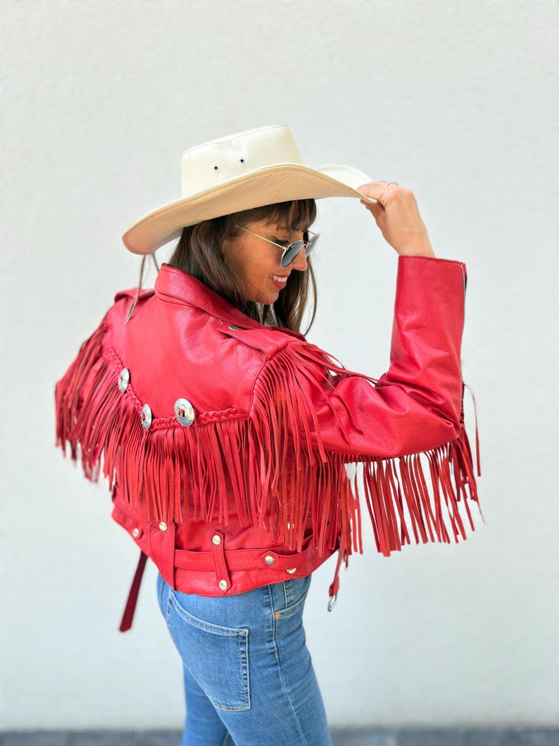 Op de afbeelding: Rode leren jas met lange franjes en zilverkleurige versieringen. De jas wordt gecombineerd met een blauwe spijkerbroek en een cr&egrave;mekleurige cowboyhoed. De franjes geven een Western-stijl. De jas heeft een riem.