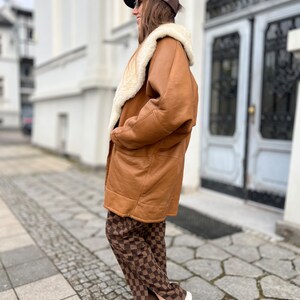 May include: A brown leather shearling coat with a white fur collar. The coat is worn over brown and white checkered pants and white sneakers.