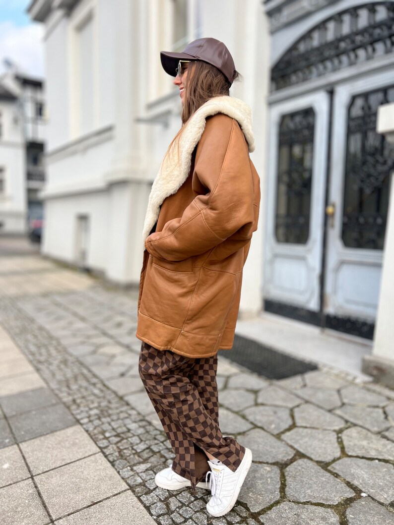 May include: A brown leather shearling coat with a white fur collar. The coat is worn over brown and white checkered pants and white sneakers.