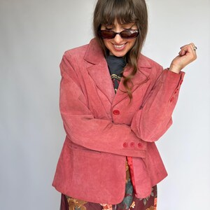 May include: A dusty rose suede blazer featuring a notched lapel, a single front button closure, and three matching buttons on each sleeve cuff. The blazer is worn over a dark graphic t-shirt and a long skirt with a floral pattern in shades of brown.