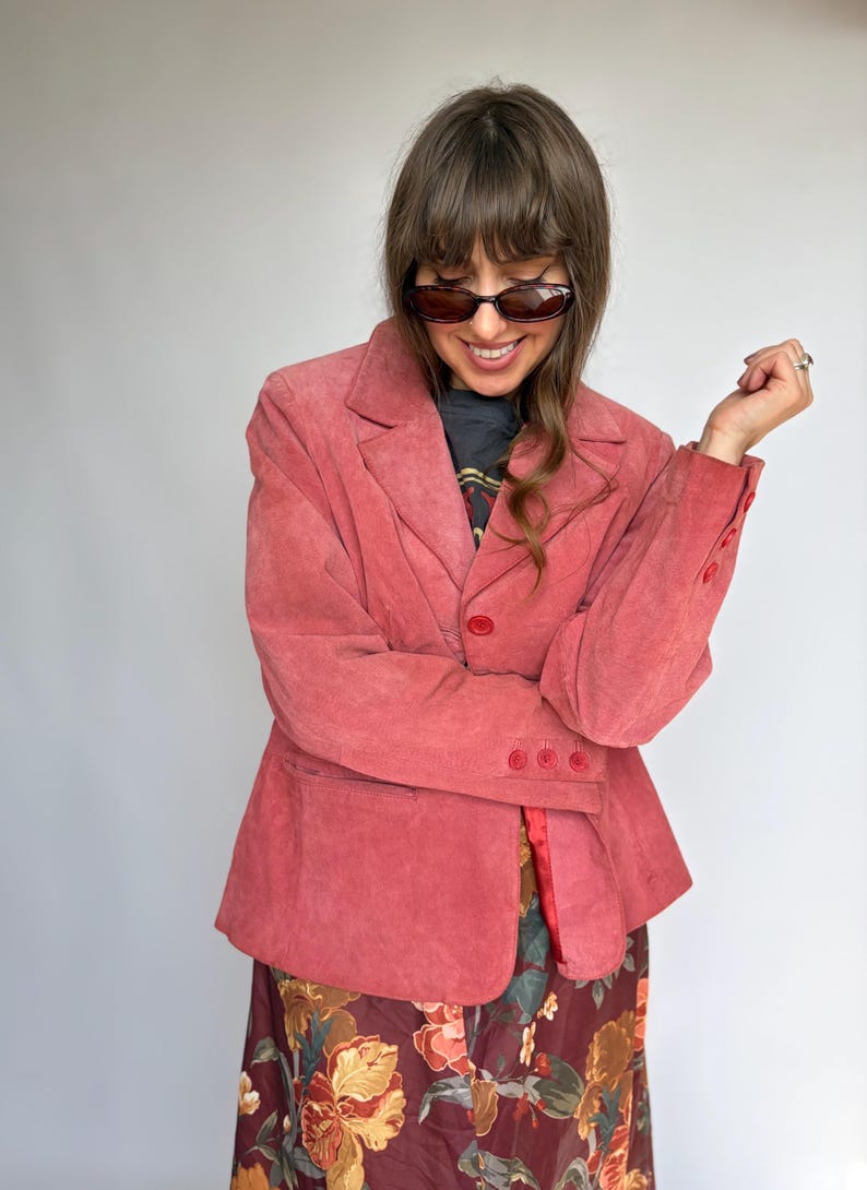 May include: A dusty rose suede blazer featuring a notched lapel, a single front button closure, and three matching buttons on each sleeve cuff. The blazer is worn over a dark graphic t-shirt and a long skirt with a floral pattern in shades of brown.