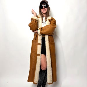 May include: A long, brown leather coat with a cream shearling lining and trim. The coat has large pockets and a button closure. The person in the image is wearing a brown cap, sunglasses, and black boots.
