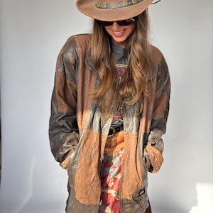 Puede incluir: Un sombrero de vaquero de cuero marrón con una banda decorativa y una chaqueta de cuero marrón con un diseño de patchwork. La chaqueta tiene paneles marrones y beige. Se ve una falda con estampado floral.