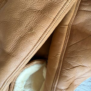 May include: Close-up of a tan leather jacket with visible stitching and a sheepskin lining. The leather has a textured appearance with subtle variations in color. The jacket's interior features a soft, cream-colored sheepskin lining.