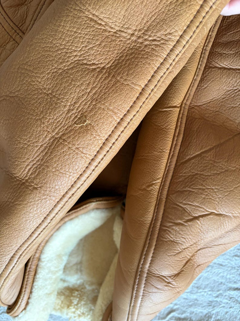 May include: Close-up of a tan leather jacket with visible stitching and a sheepskin lining. The leather has a textured appearance with subtle variations in color. The jacket's interior features a soft, cream-colored sheepskin lining.